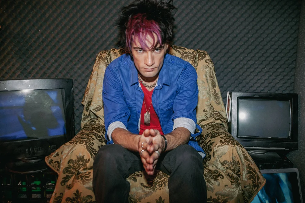 Man with colorful hair sitting on a patterned chair, surrounded by TV screens with a soundproof background.