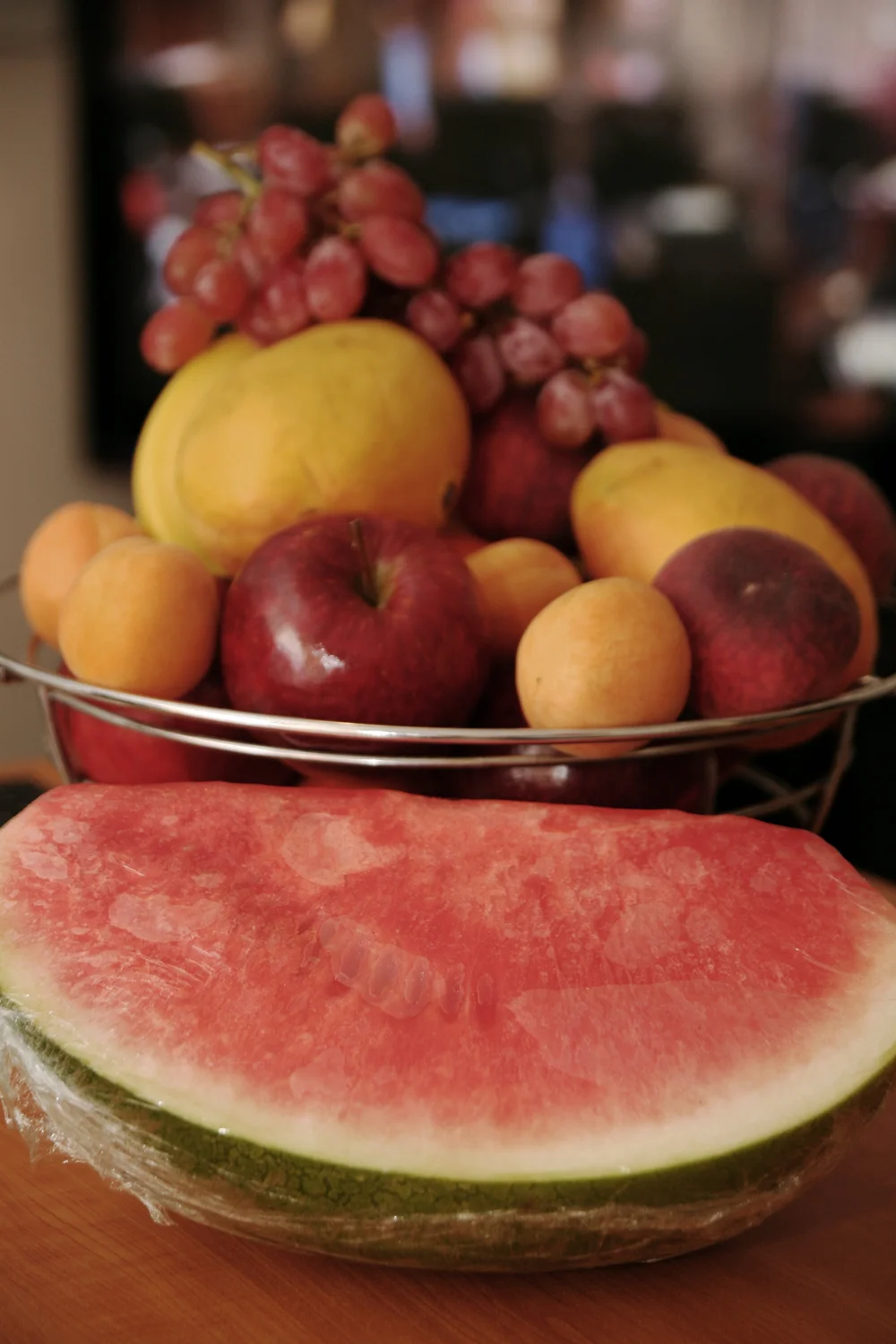 A variety of fruits including a large slice of watermelon, apples, grapes, peaches, and apricots in a wire basket.