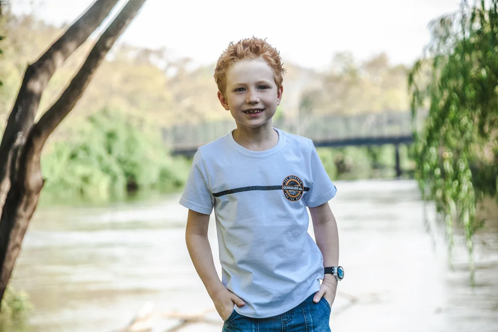 Smiling boy wearing a white T-shirt and jeans in front of a river with trees and a bridge in the background.