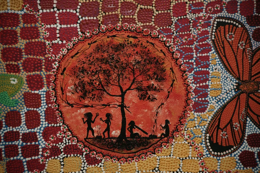 Aboriginal art depicting silhouettes of people around a tree, vibrant dot painting background, butterfly on the side.