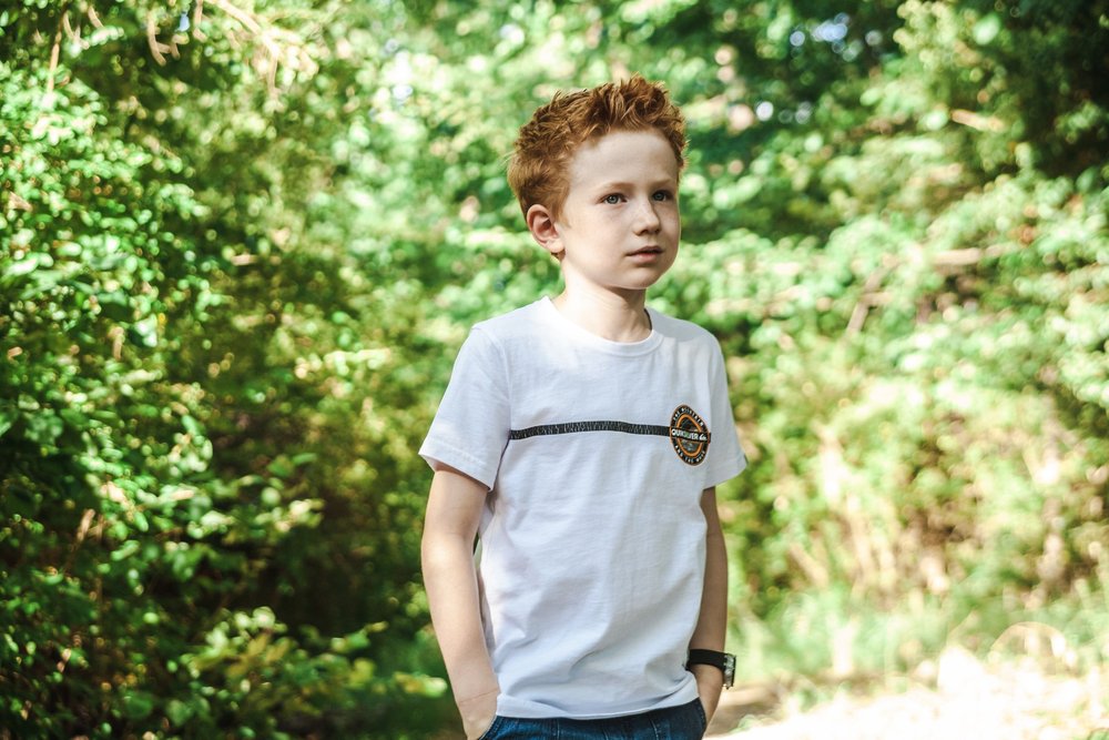 Young boy with red hair in white shirt standing outdoors in a forested area.