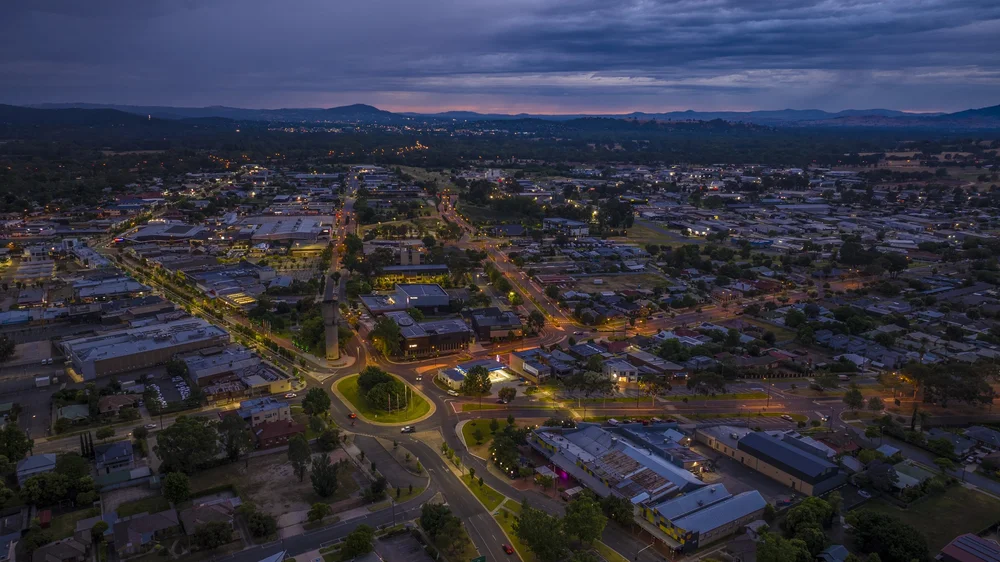 Aerial view of a town during twilight, showing illuminated streets, buildings, and a roundabout, surrounded by trees and distant hills under a cloudy sky.