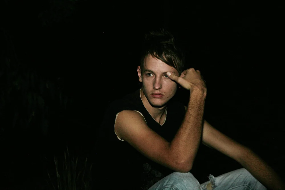 Person sitting at night in a thoughtful pose, hand on face, wearing a sleeveless shirt and jeans; dark background.