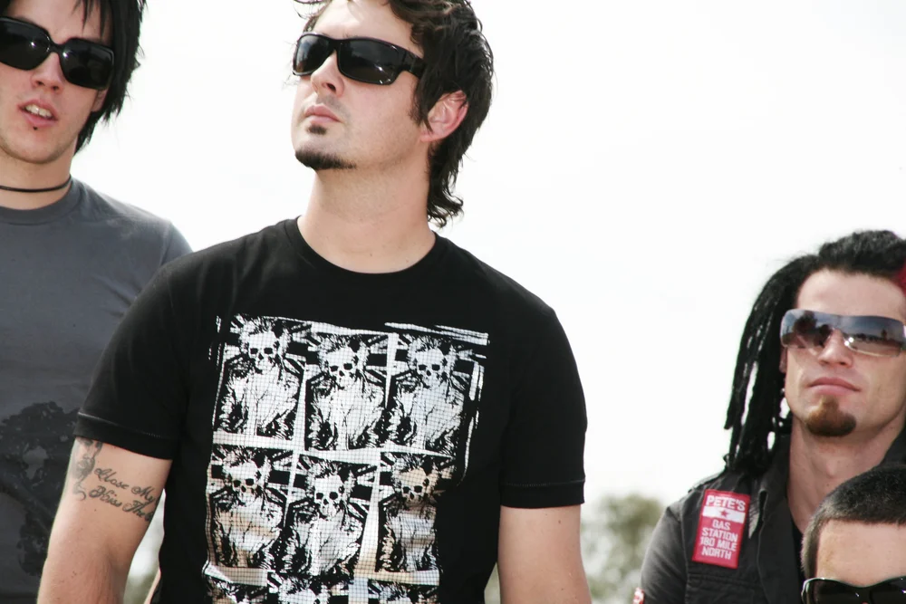 Three men wearing sunglasses, one in a skull print shirt, one with dreadlocks, outdoor setting.