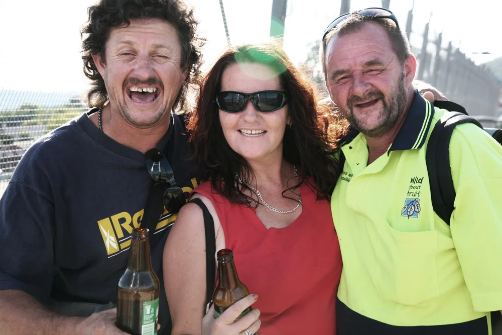 Group of three people outdoors, smiling and holding beer bottles, two men and one woman wearing casual clothing.