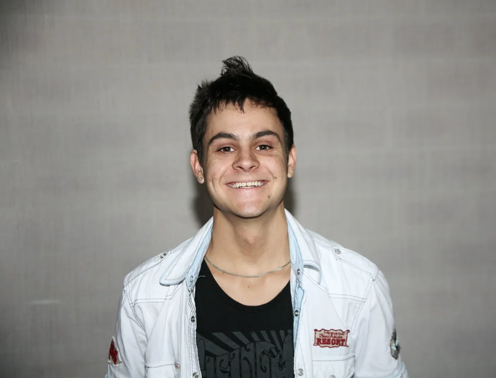 Young man smiling against a plain background, wearing a light-colored jacket over a black shirt.