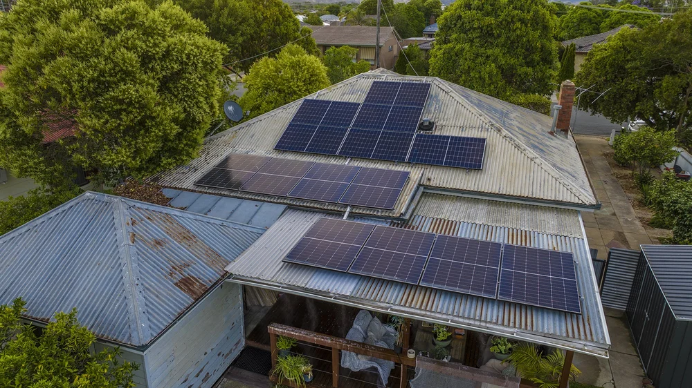 Aerial view of a house with multiple solar panels on a corrugated metal roof, surrounded by trees and greenery.