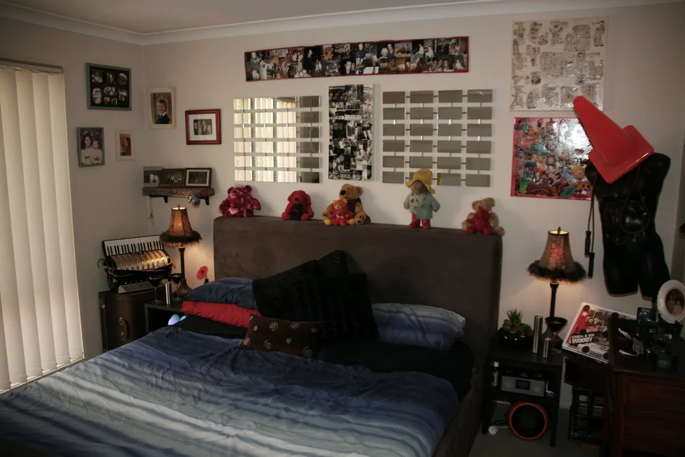 Bedroom with double bed, pillows, and plush toys on headboard. Walls decorated with photos, mirrors, artwork, and mannequin with traffic cone. Accordion on side table.
