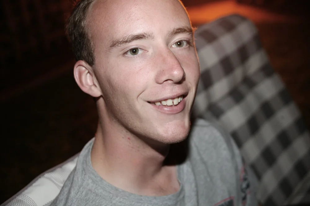 Close-up portrait of a smiling person outdoors at night, wearing a gray shirt, with a plaid-patterned background.