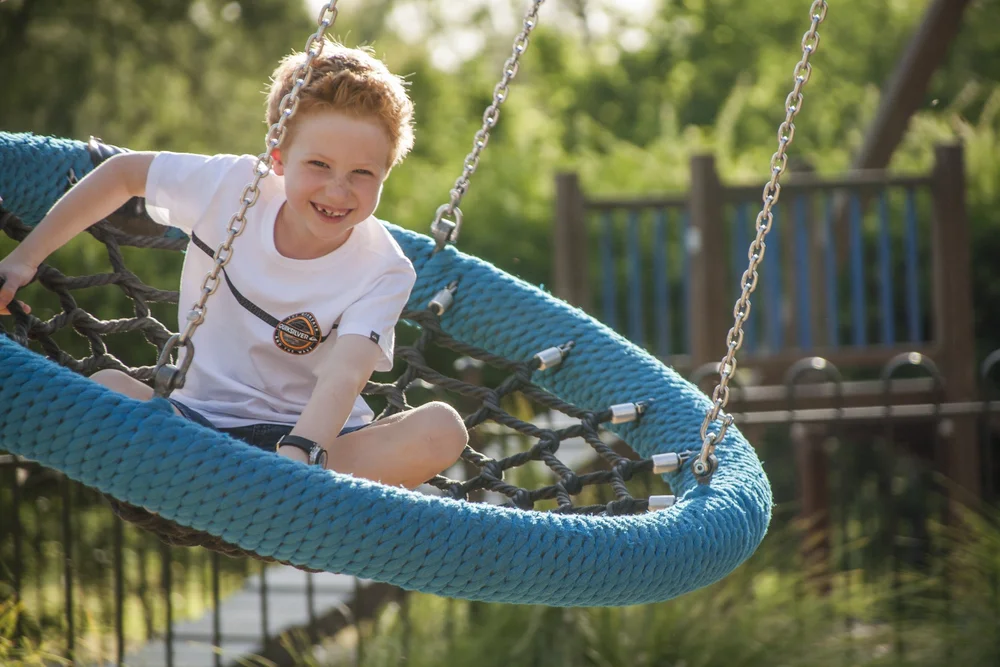 Child smiling on a blue net swing in a playground.