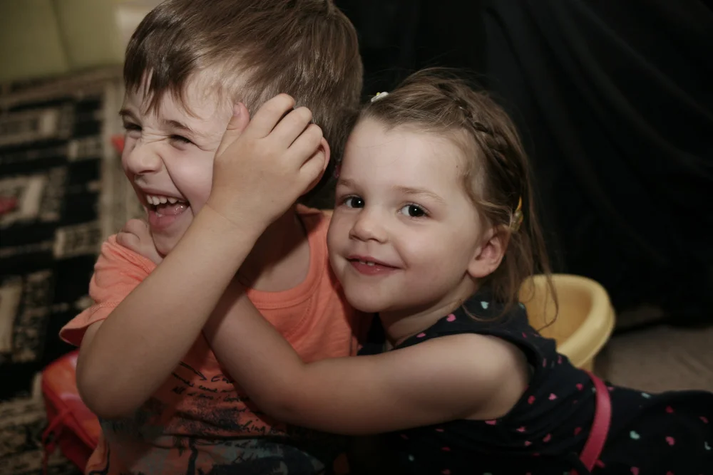 Two young children laughing and hugging. The boy is wearing an orange shirt, and the girl is in a dark dress with colorful dots.