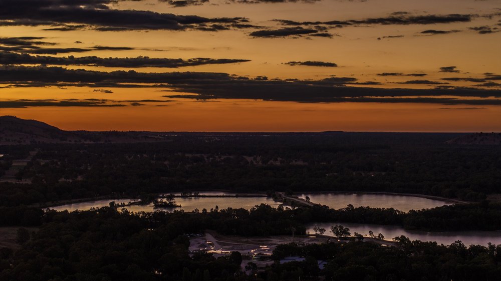 Aerial view of a serene lake surrounded by dense forest during sunset, with an orange and purple sky.