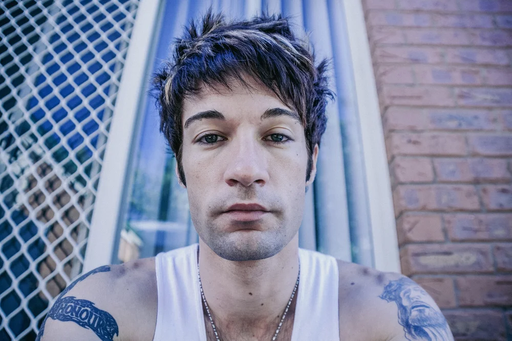 Man with spiky hair and tattoos sitting in front of a screen door and brick wall, wearing a tank top.