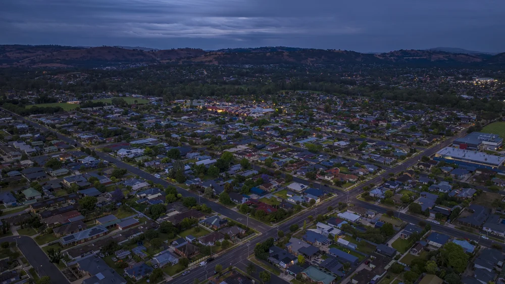 Aerial view of a suburban residential area at dusk, with rows of houses, streets, and scattered streetlights, surrounded by hills in the background under a cloudy sky.