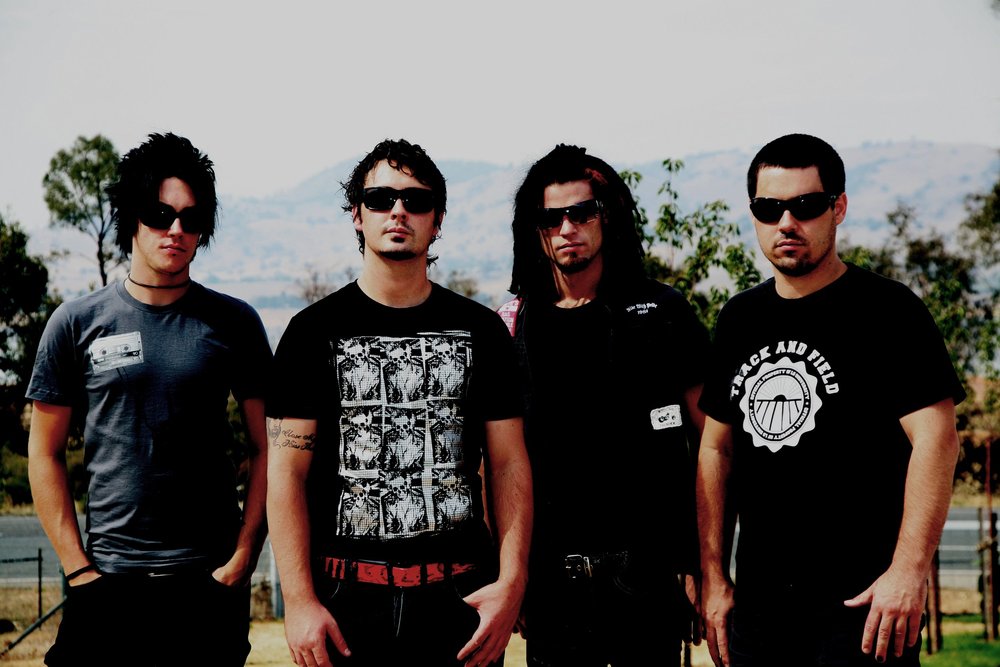 Four men wearing sunglasses standing outdoors with trees and a mountainous background.