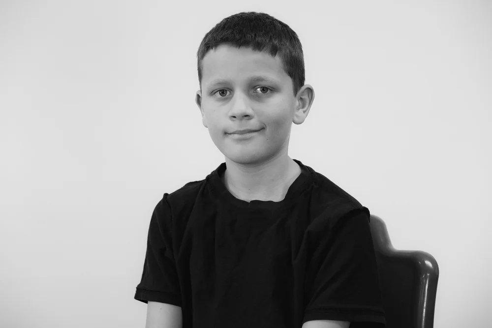 Black and white portrait of a boy sitting and smiling, wearing a dark T-shirt, with a plain background.