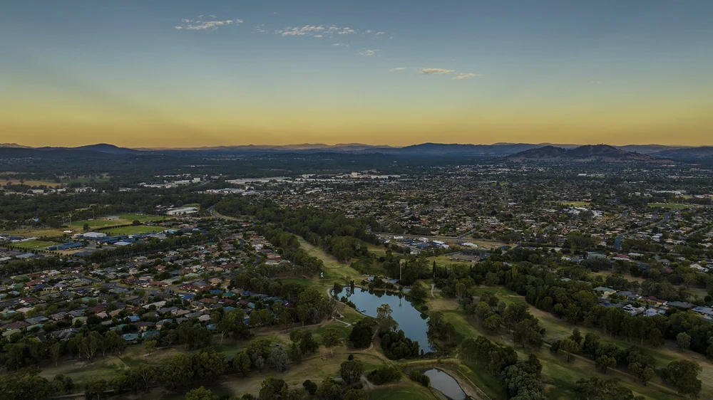Aerial view of Wagga Wagga with residential areas, green spaces, and distant mountains under a sunset sky.