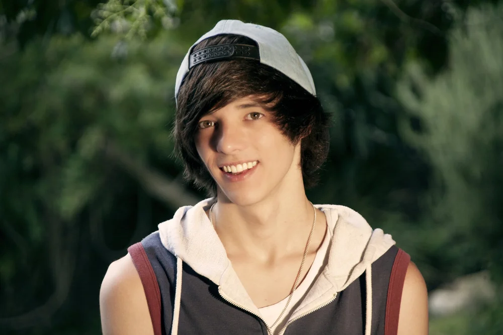 Young person smiling, wearing a cap backwards and a sleeveless hoodie, outdoors with greenery in the background.