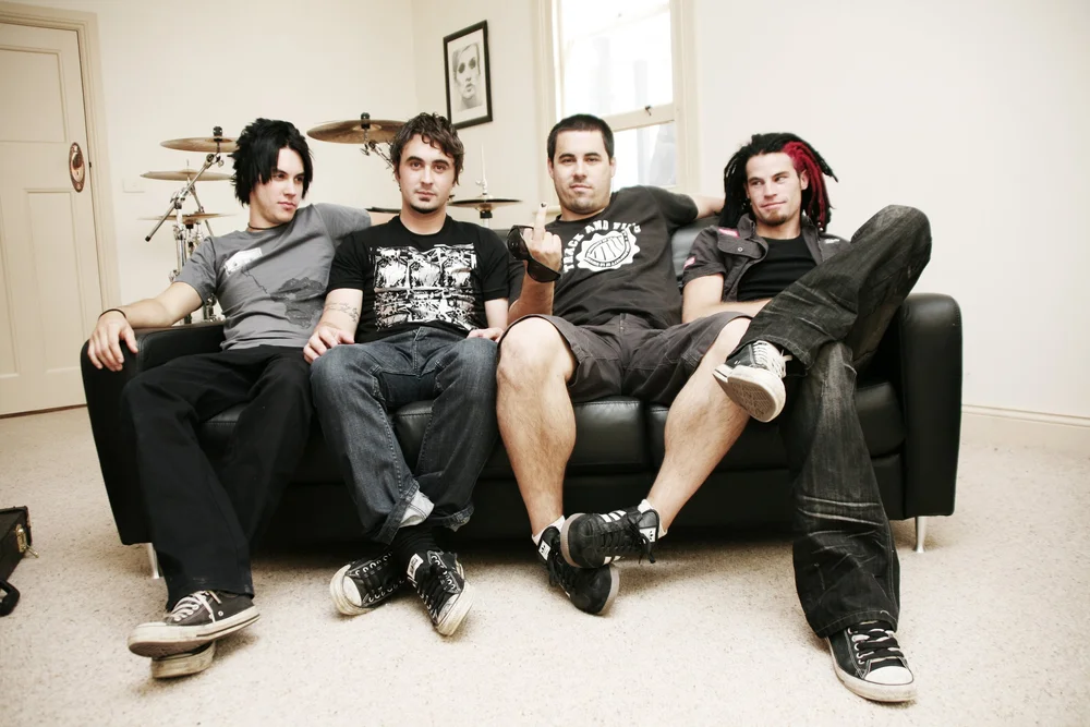 Four men sitting on a black sofa in a room with a drum set in the background.