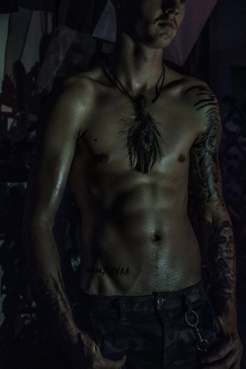 Shirtless man with tattoos and feather necklace in dim lighting