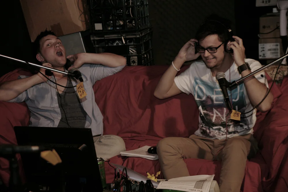 Two men sitting on a red couch with microphones, wearing headphones in a recording studio.