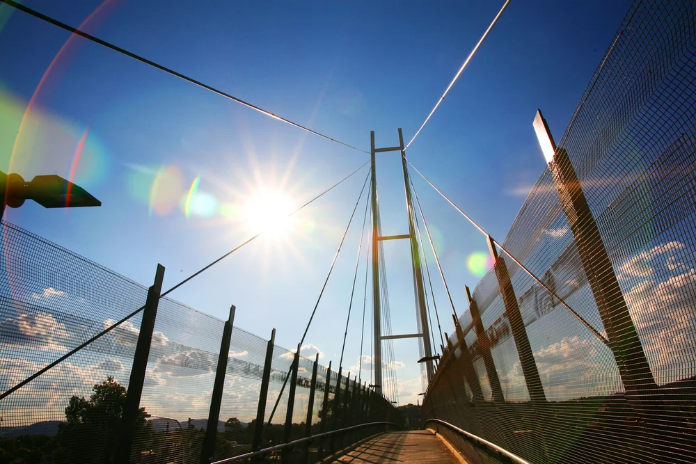 Sunlit pedestrian bridge with metal fencing and cables against blue sky