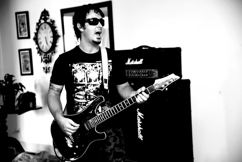 Black and white photo of a musician playing an electric guitar with a Marshall amplifier, wearing sunglasses and a graphic t-shirt.