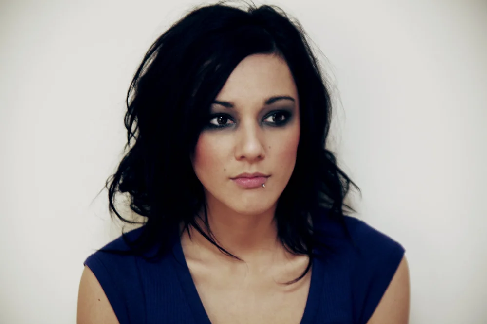 Woman with dark hair, wearing a blue top, and a lip piercing, looking away from the camera.