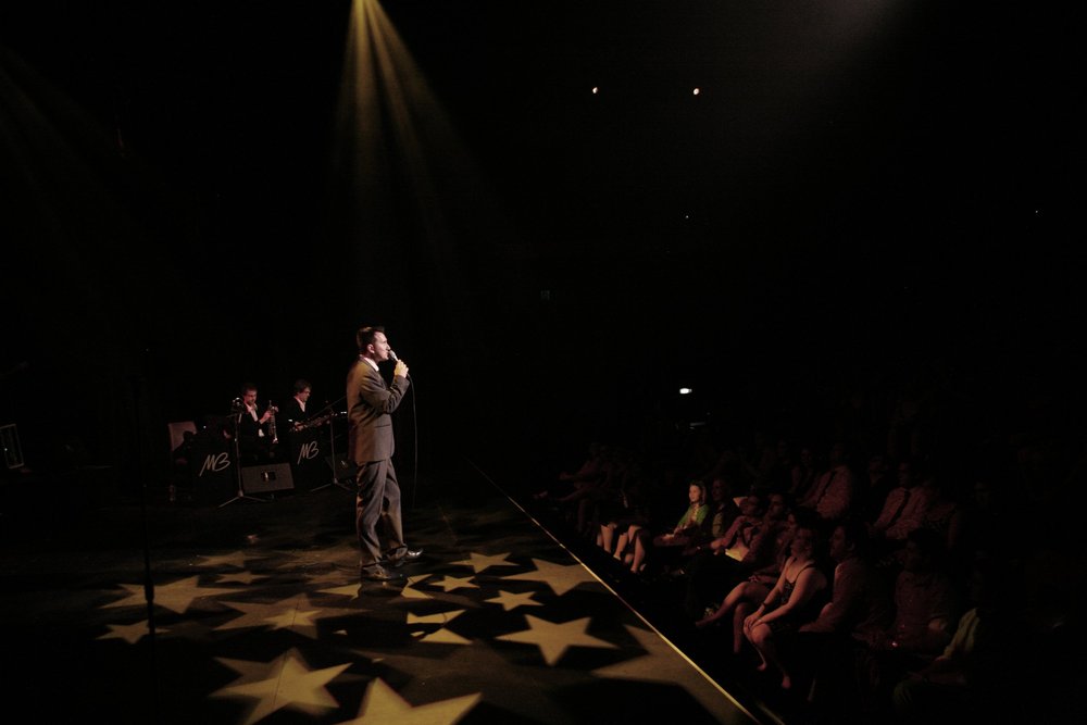 Singer performing on stage with spotlight, accompanied by a band, and audience watching.