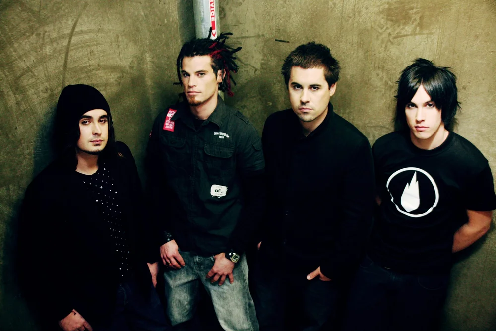 Four men standing against a concrete wall, dressed in dark casual clothing.