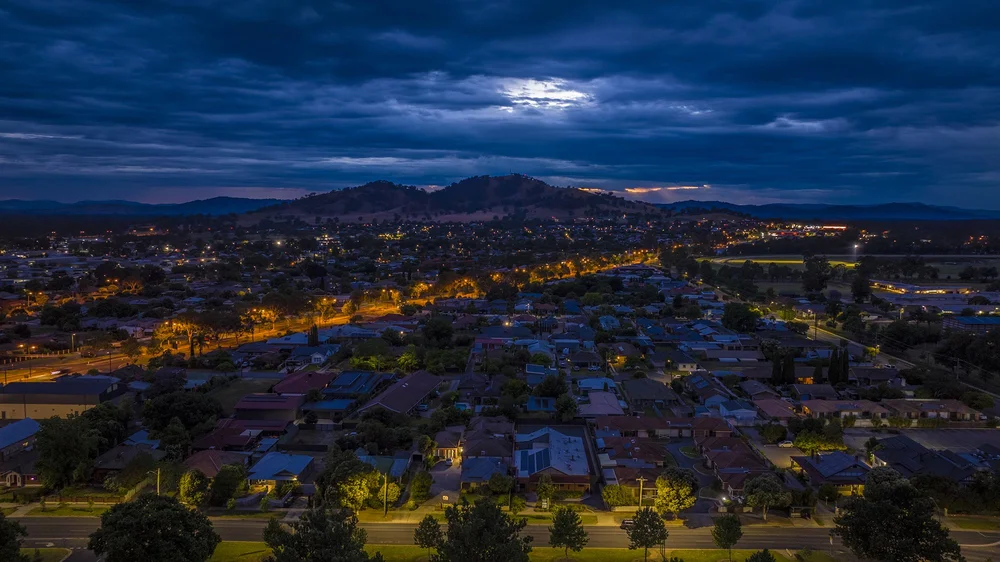 Aerial view of a town at dusk with illuminated streets and houses, surrounded by hills and mountains under a cloudy sky.