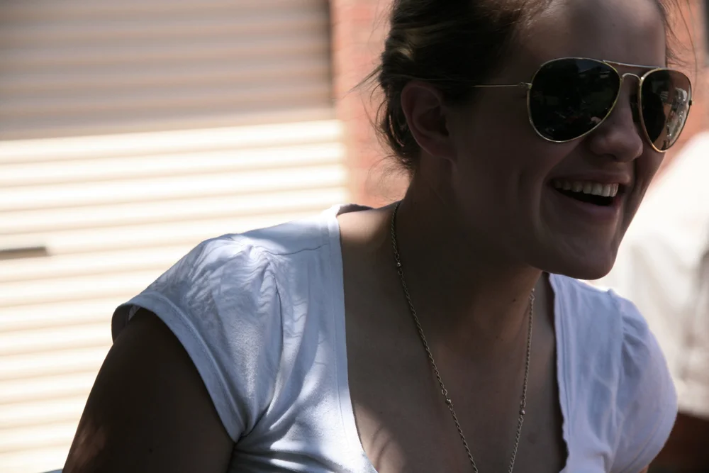 Person smiling, wearing sunglasses, white shirt, and necklace with blurred background.