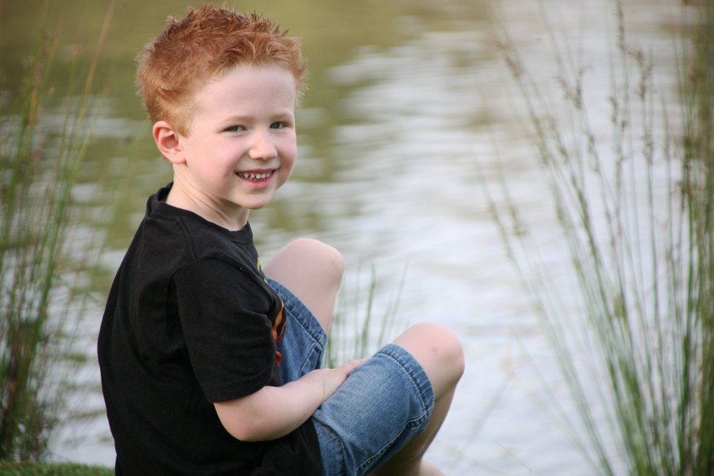 Young boy sitting by a pond, wearing black shirt and denim shorts, smiling.