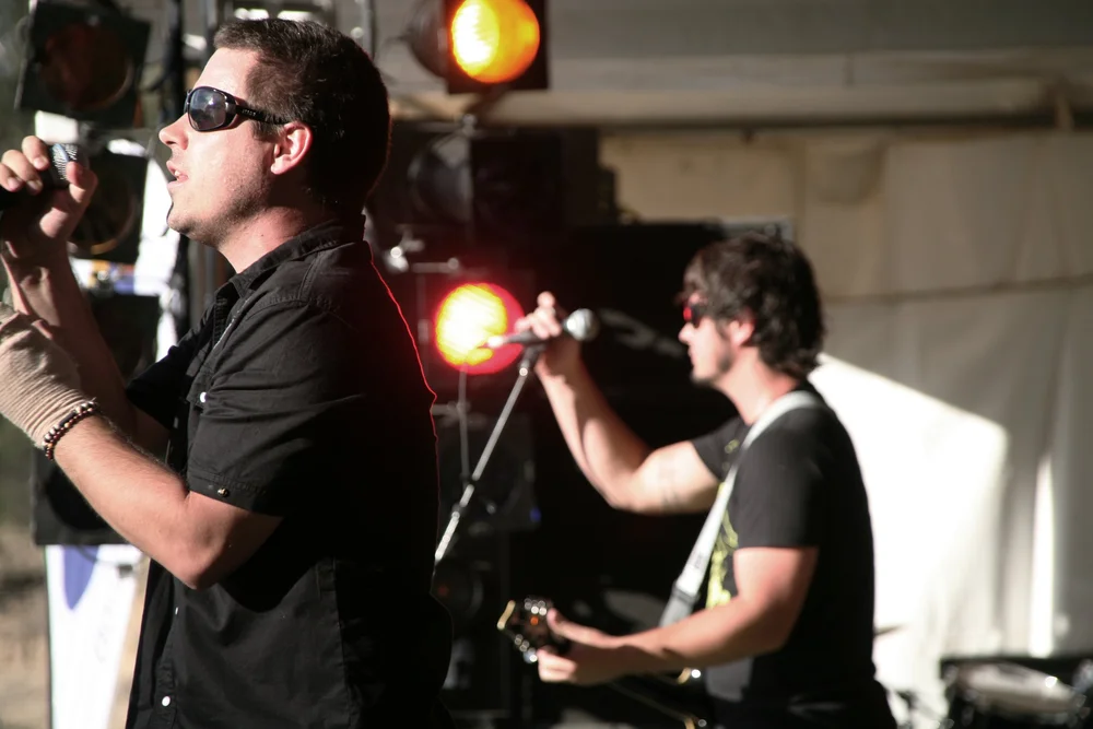 Musicians performing on stage with microphones and instruments, wearing sunglasses, with stage lights in the background.
