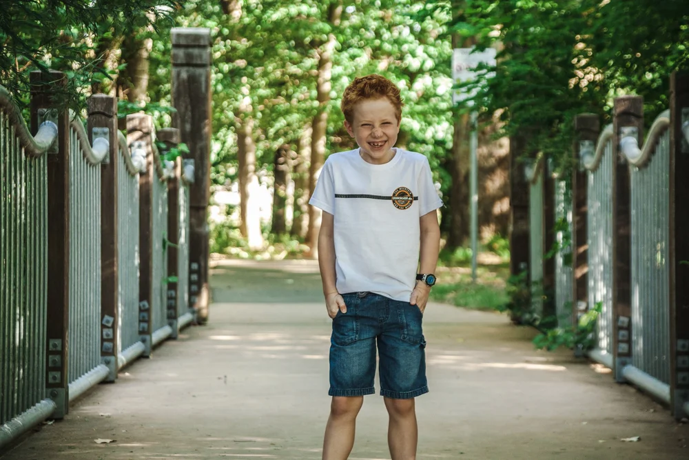 Smiling child on a park walkway wearing a white t-shirt and denim shorts, surrounded by greenery.