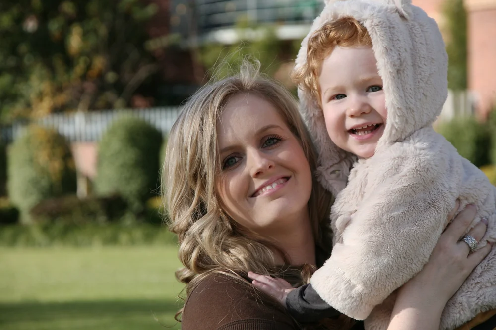 Smiling woman holding a young child in a bear hoodie outdoors.