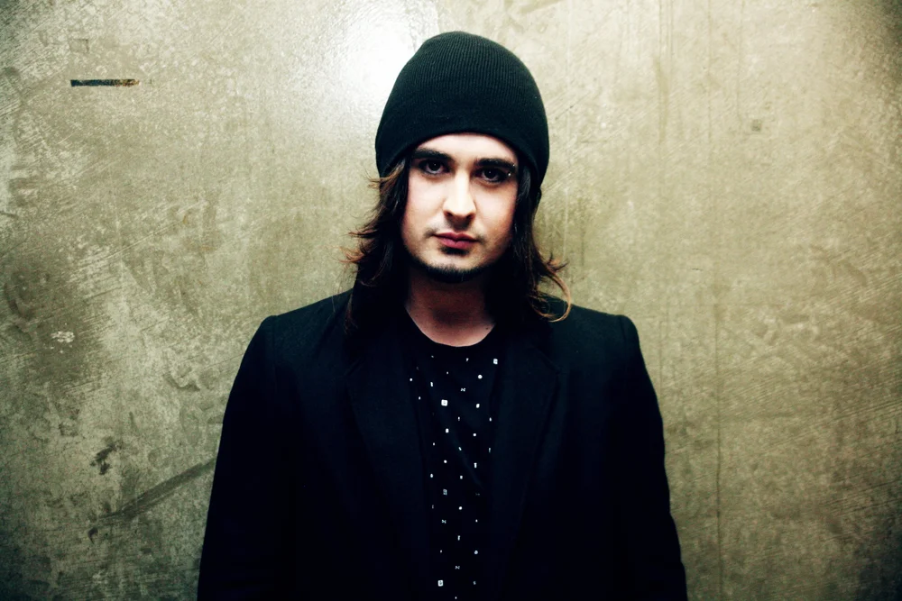 Person wearing a black beanie and black jacket standing against a textured wall.