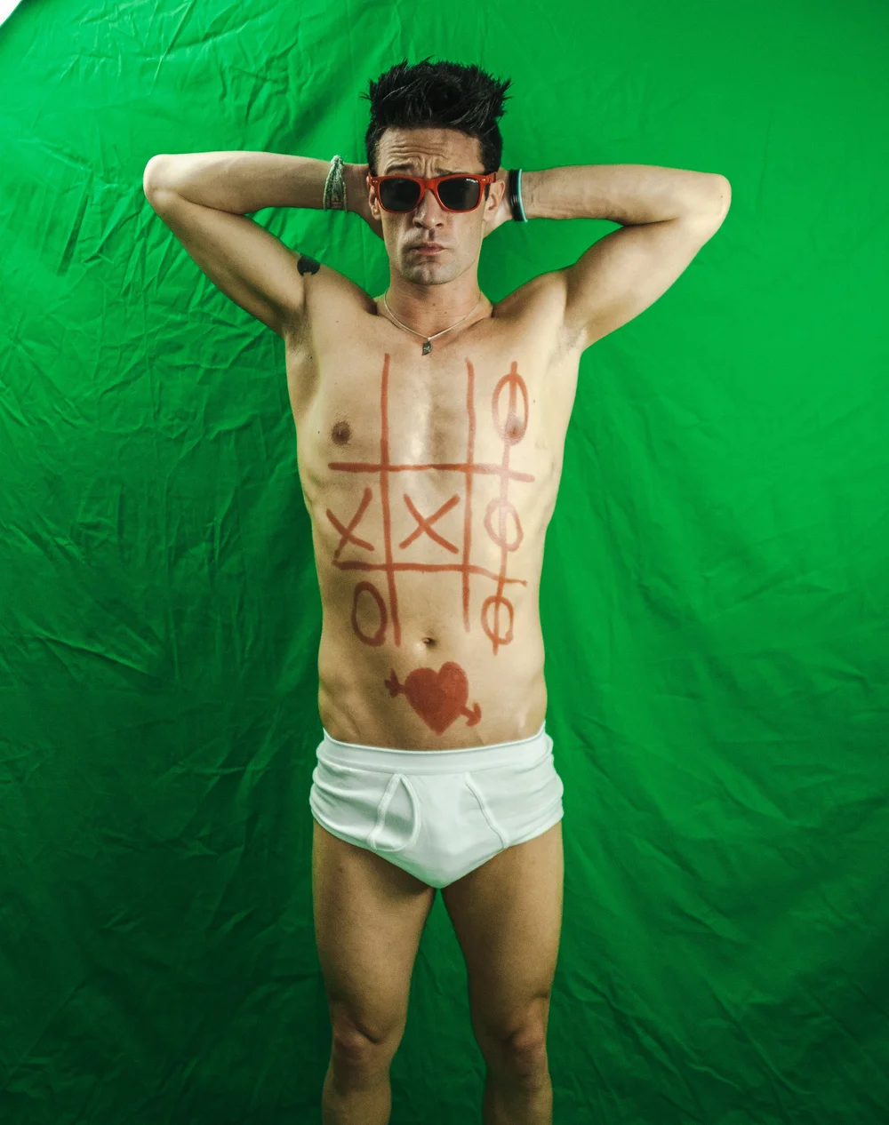 Man with tic-tac-toe drawn on torso, wearing red sunglasses and white underwear, green background.