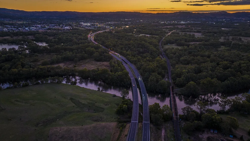Aerial view of a highway winding through a lush forest landscape with a river, during sunset.