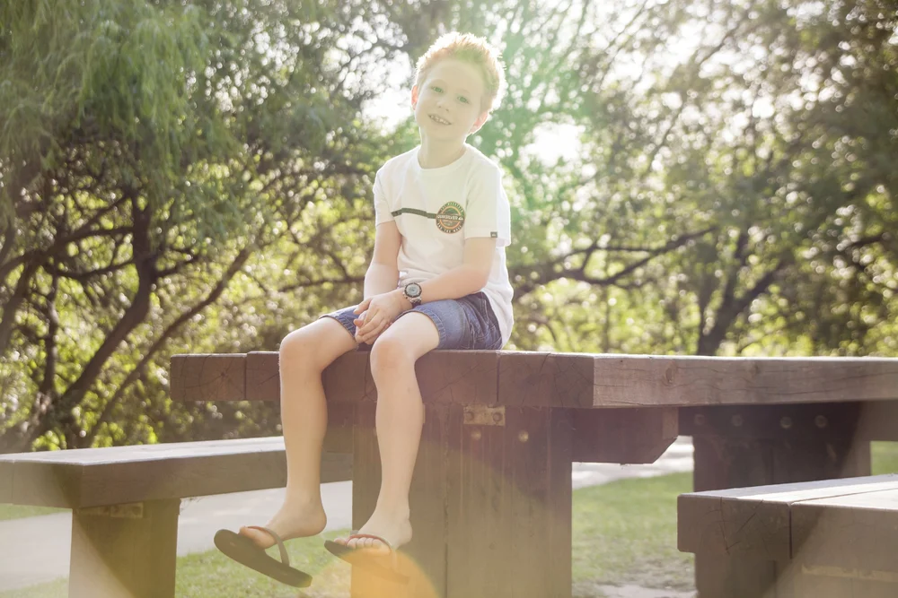 Child sitting on a wooden bench in a park, wearing a white shirt and shorts, surrounded by trees and sunlight.