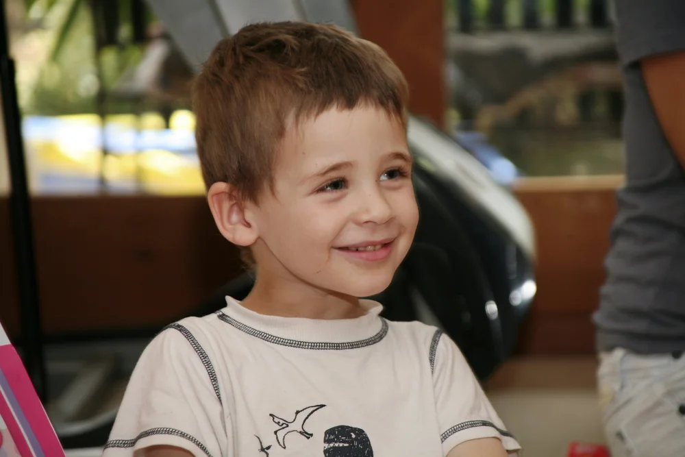 Young child smiling indoors, wearing a light-colored shirt with graphic design, blurred background.