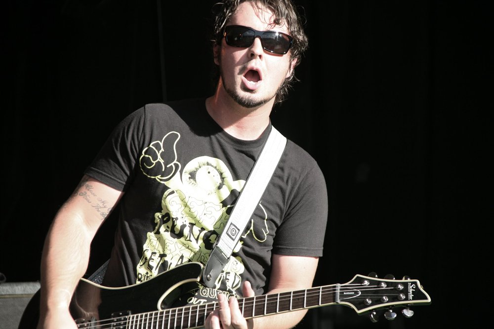 Musician playing guitar wearing sunglasses and a black t-shirt with a graphic print.