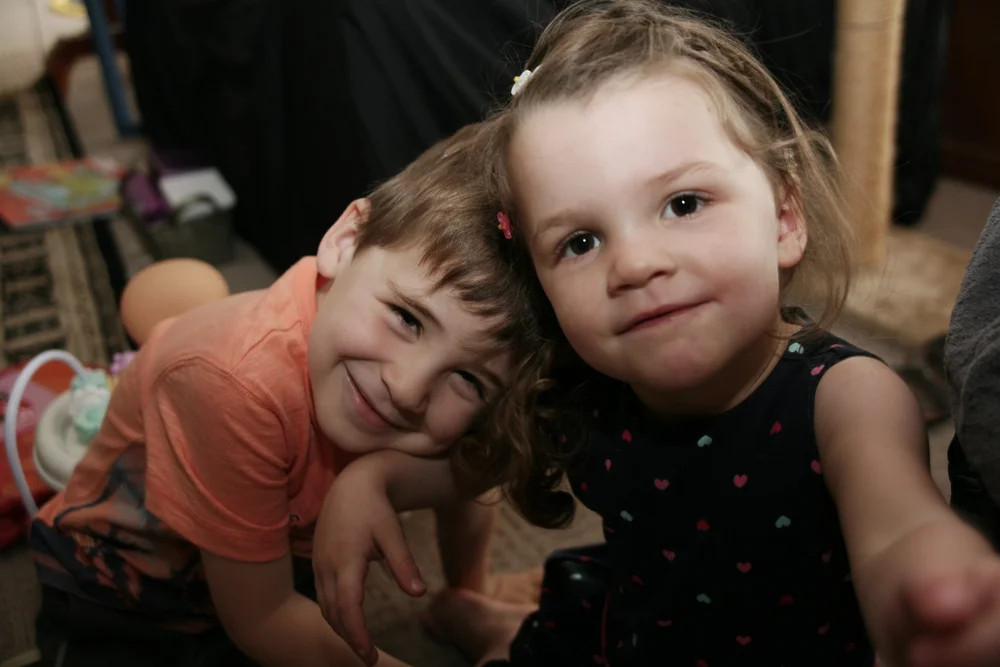 Two young children smiling indoors, one wearing an orange shirt, the other in a dark dress with polka dots.