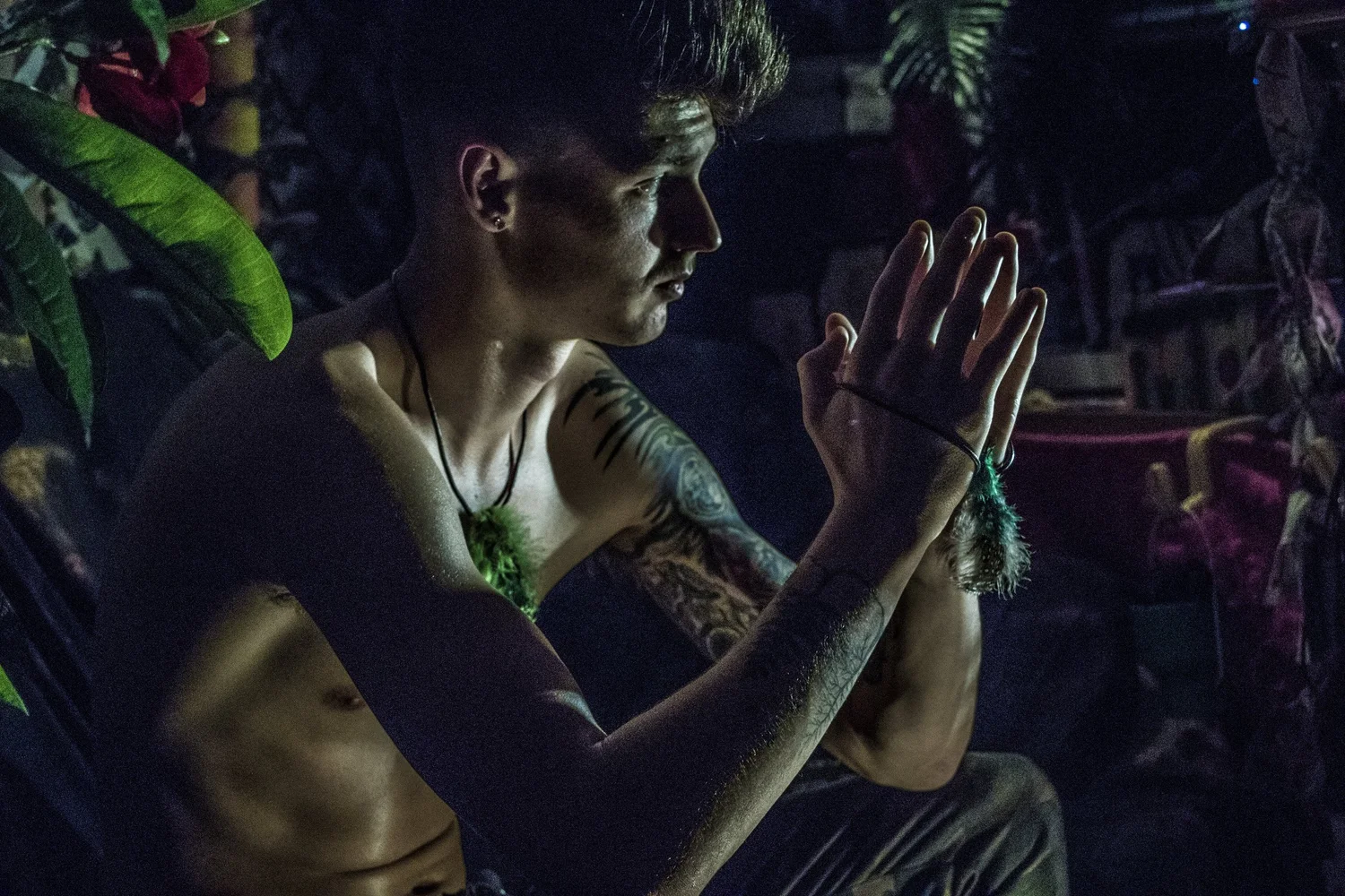 Low-light portrait by Amos Ashley: a tattooed person in profile with hands held together, surrounded by layered colour and objects.