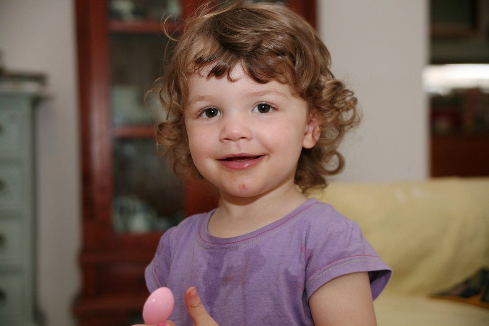 Young child with curly hair smiling, holding a pink object, wearing a purple shirt, indoors.