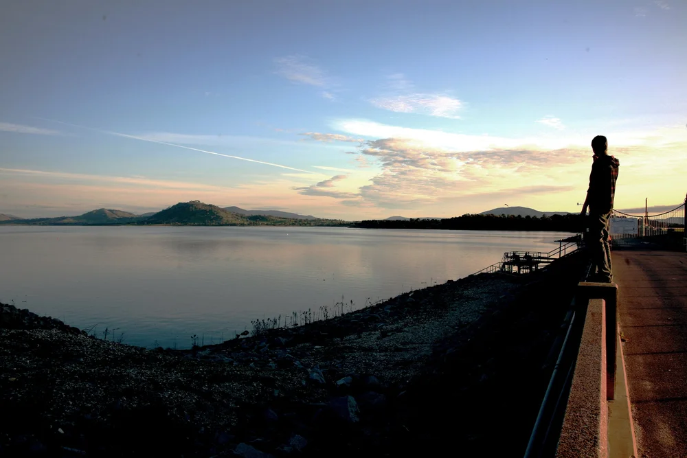 Silhouette of a person standing on a railing by a calm lake with hills in the background at sunset.