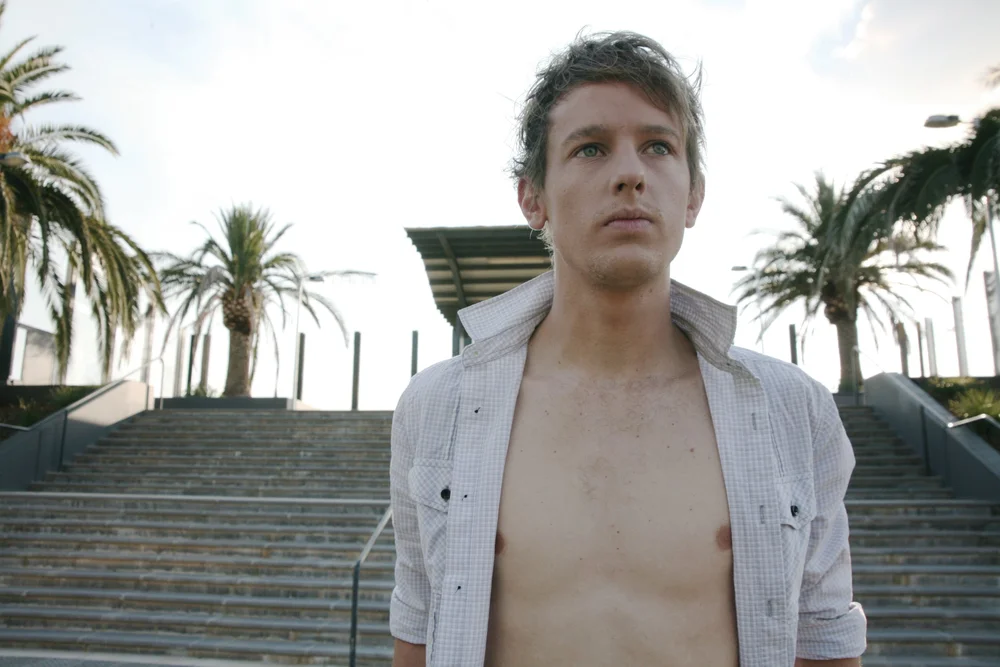 Man standing shirtless in front of outdoor staircase with palm trees.