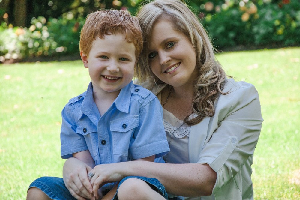 A woman and young boy smiling outdoors, with greenery in the background.