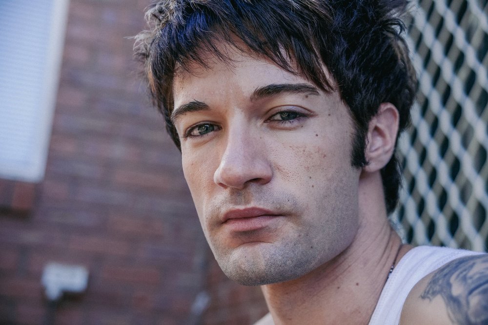 Close-up of a man with dark hair and facial stubble, wearing a white tank top, with a blurred brick wall background.