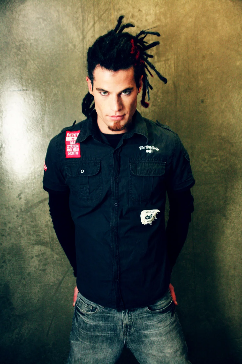 Man with dreadlocks wearing a dark shirt and jeans against a textured wall.